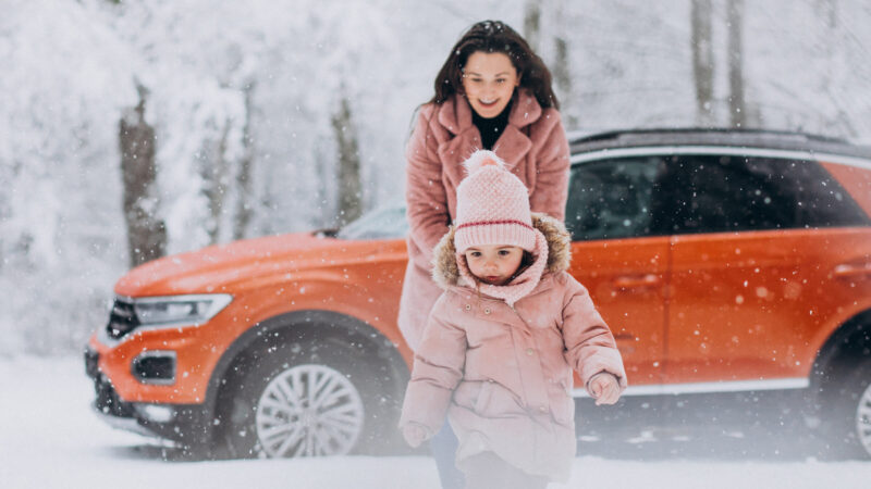 How to prepare your car for winter - Cover Photo | Warrantywise A woman and a young child dressed in warm clothes play in the snow near an orange car.
