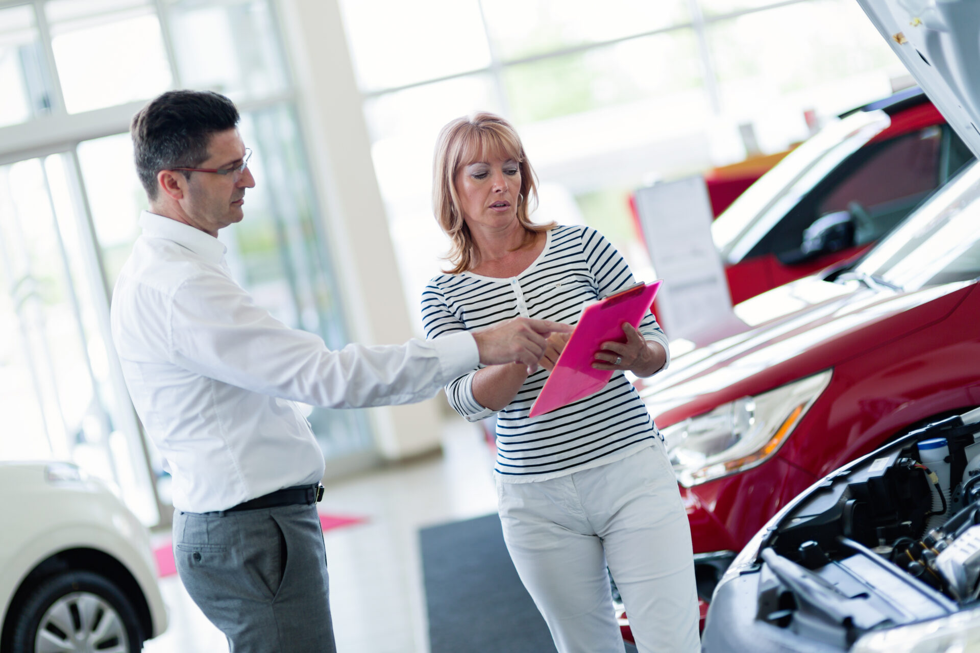 auto business car sale consumerism and people concept | Warrantywise Woman discussing car details with a salesperson at a dealership, reviewing paperwork near a vehicle.
