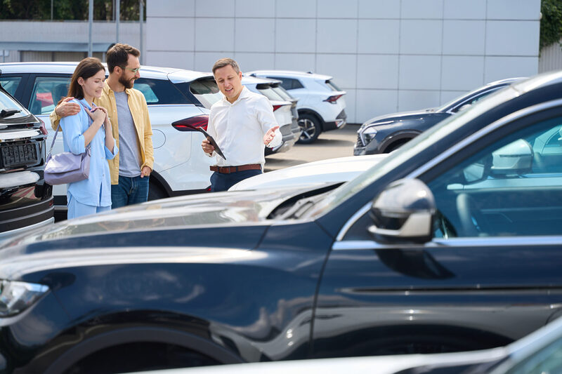 Buyers choose a car in the yard of car dealership | Warrantywise A car salesperson showing a couple different vehicles at a dealership, discussing features and options for buying a car.