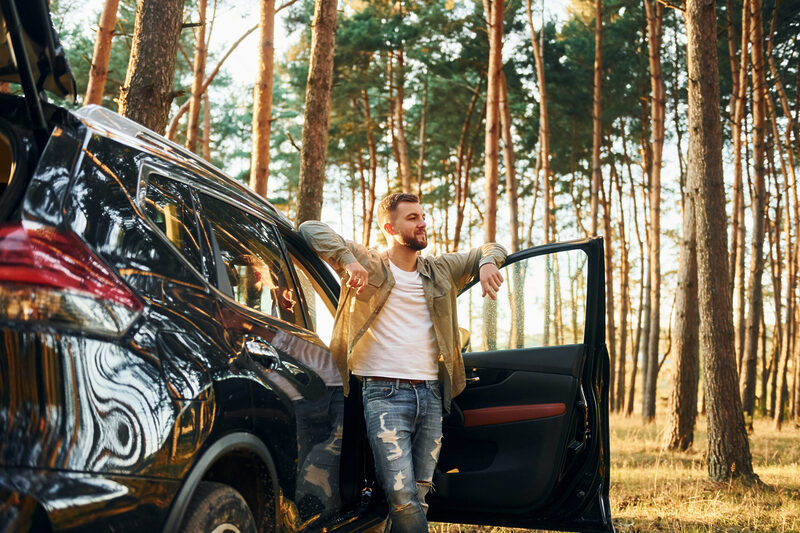 A man leans against an open car door in a forest, looking out while sunlight filters through the trees