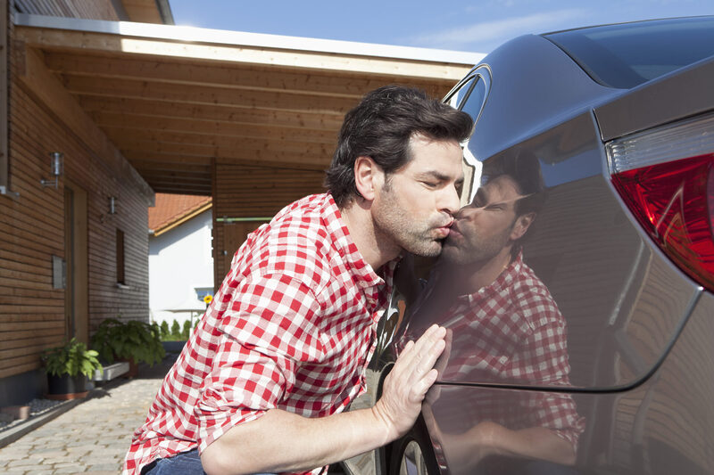 A man in a red check shirt kisses the side of a car, with his reflection visible in the shiny paint