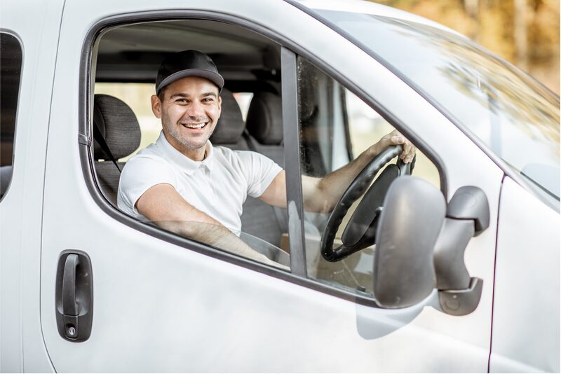 A smiling van driver sits behind the wheel of a white van, looking out through the open window