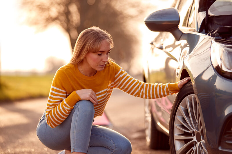 Woman Broken Down On Country Road Checking Car Tyre For Puncture | Warrantywise Person inspecting a car tyre on a roadside, checking tread and condition as part of regular vehicle maintenance.