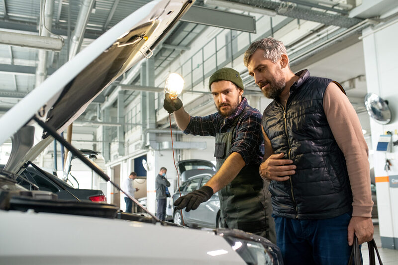 Two men bending over car engine while technician pointing at one of its parts | Warrantywise A mechanic and a customer stand beside an open car hood inside a modern auto repair workshop. The mechanic holds a work light while explaining something to the customer, who looks attentive.