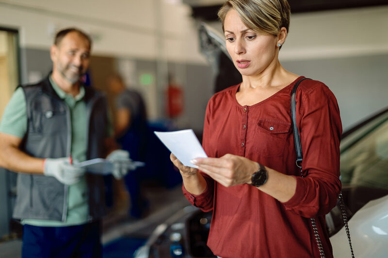 Mid adult woman feeling shocked with expense of car repairing in a workshop | Warrantywise Woman looking concerned as she reads a piece of paper in a garage, with a mechanic holding paperwork and a car nearby