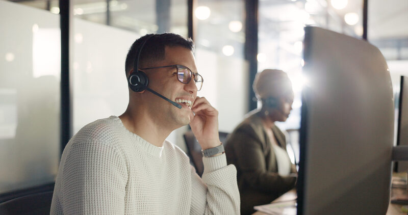 contact-us-crm-or-happy-consultant-in-call-center-2026-01-09-09-40-07-utc | Warrantywise Customer service agent wearing a headset smiles while speaking at a computer desk in a modern office, with another agent working in the background